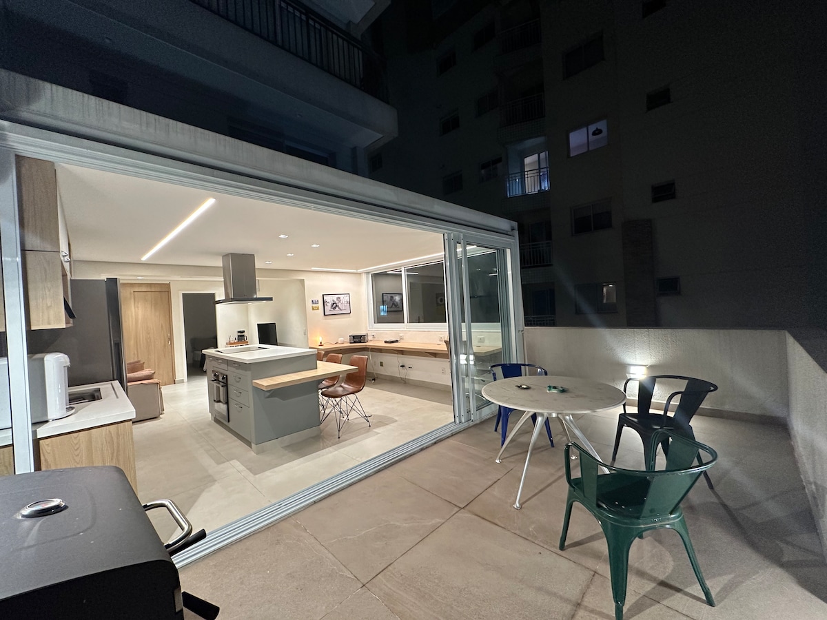 An outdoor terrace is highlighted, featuring a circular dining table accompanied by two green chairs. A barbecue grill stands nearby. The spacious interior kitchen and living area are visible through large sliding glass doors, illuminated and welcoming, contributing to a seamless indoor-outdoor flow.