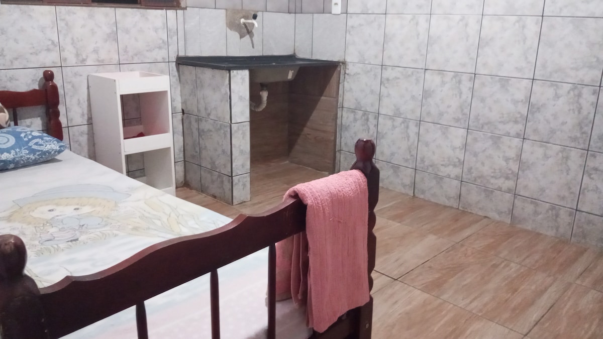 A simple individual bedroom features a wooden bed with a decorative pillow and a soft blanket. A small white shelf stands beside the bed. A compact wash area, with a sink, is positioned against a tiled wall, enhancing the room's functionality.