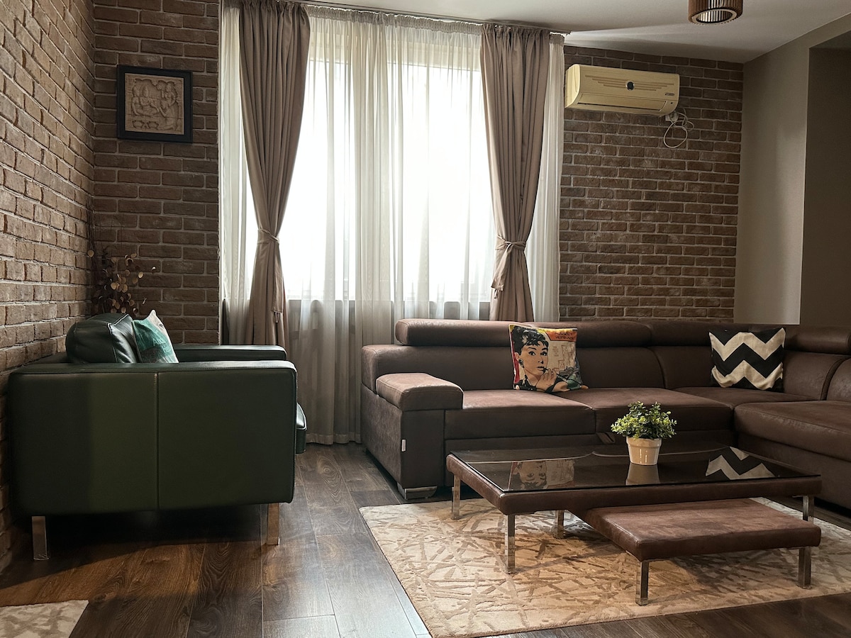 A modern living area features a rich brown sectional sofa and a green armchair, complemented by a glass coffee table atop a patterned area rug. Large windows with light curtains allow natural light to fill the space, while exposed brick walls add texture to the room.