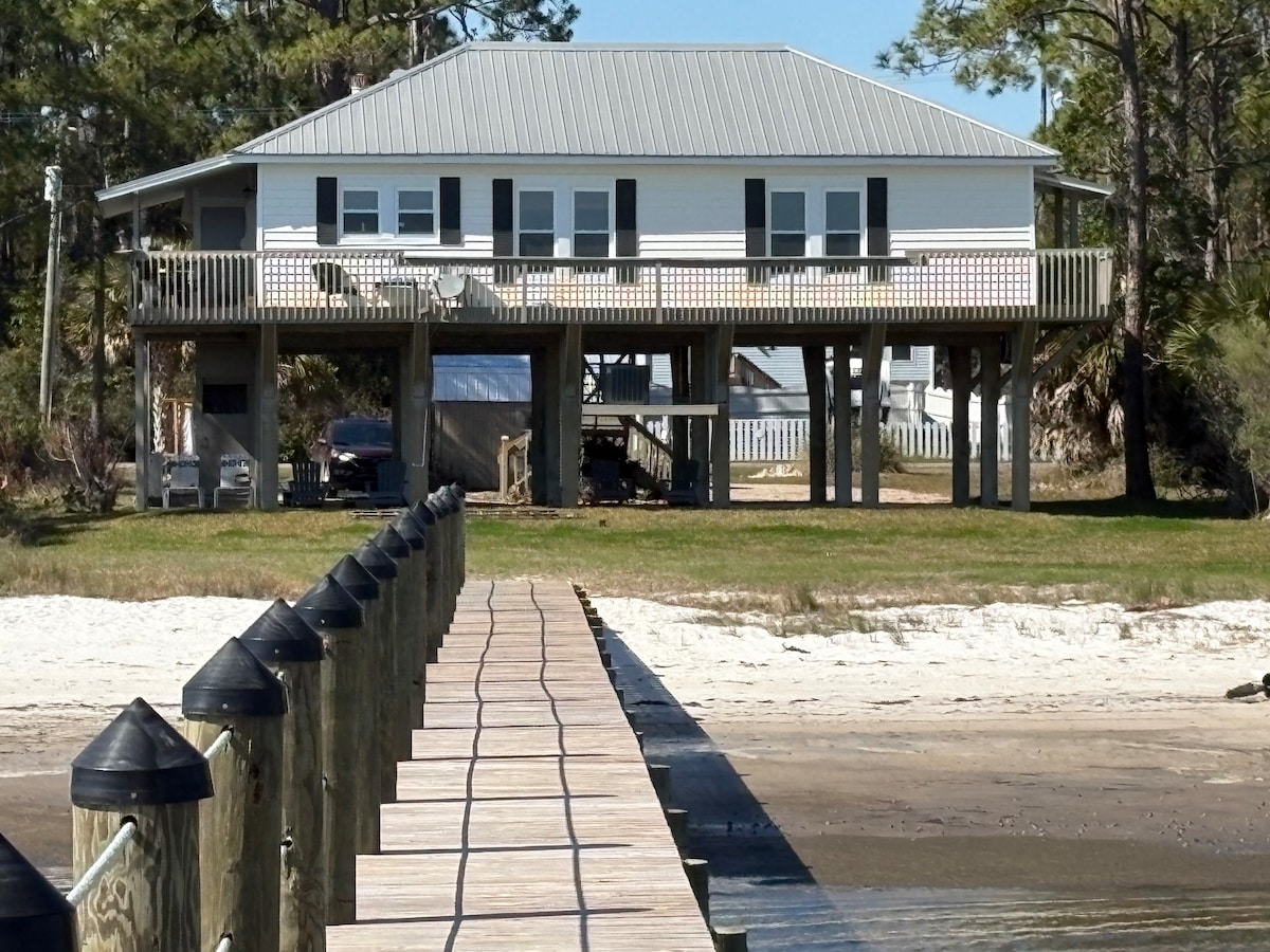 A charming waterfront farmhouse is elevated on pilings, featuring a spacious deck with black shutters on windows. The surrounding yard is adorned with lush grass, and a wooden pier leads to the sandy beach, offering direct access to the bay.