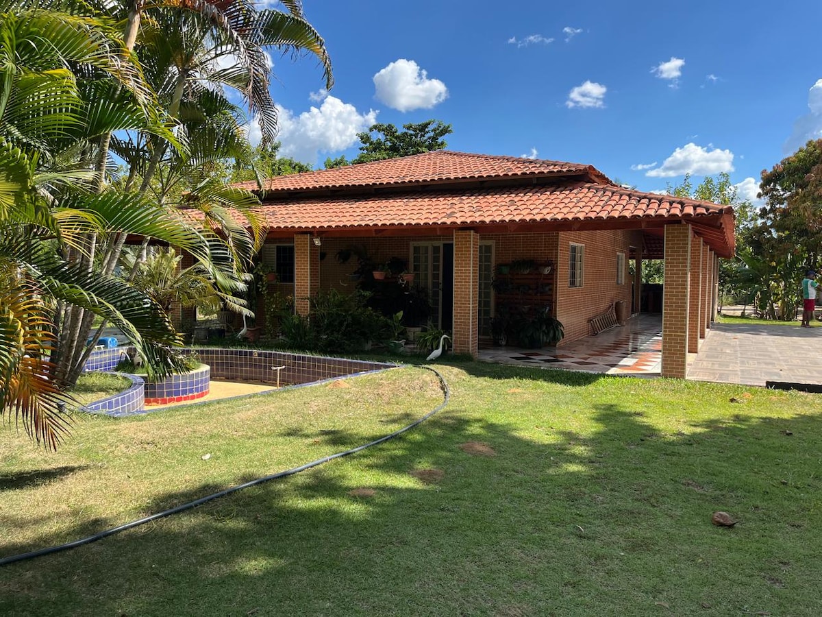 The image displays a charming brick house surrounded by a green lawn and palm trees. A covered porch offers shaded seating. A swimming pool is partially visible, framed by colorful tiles. The clear blue sky creates a bright and inviting atmosphere.