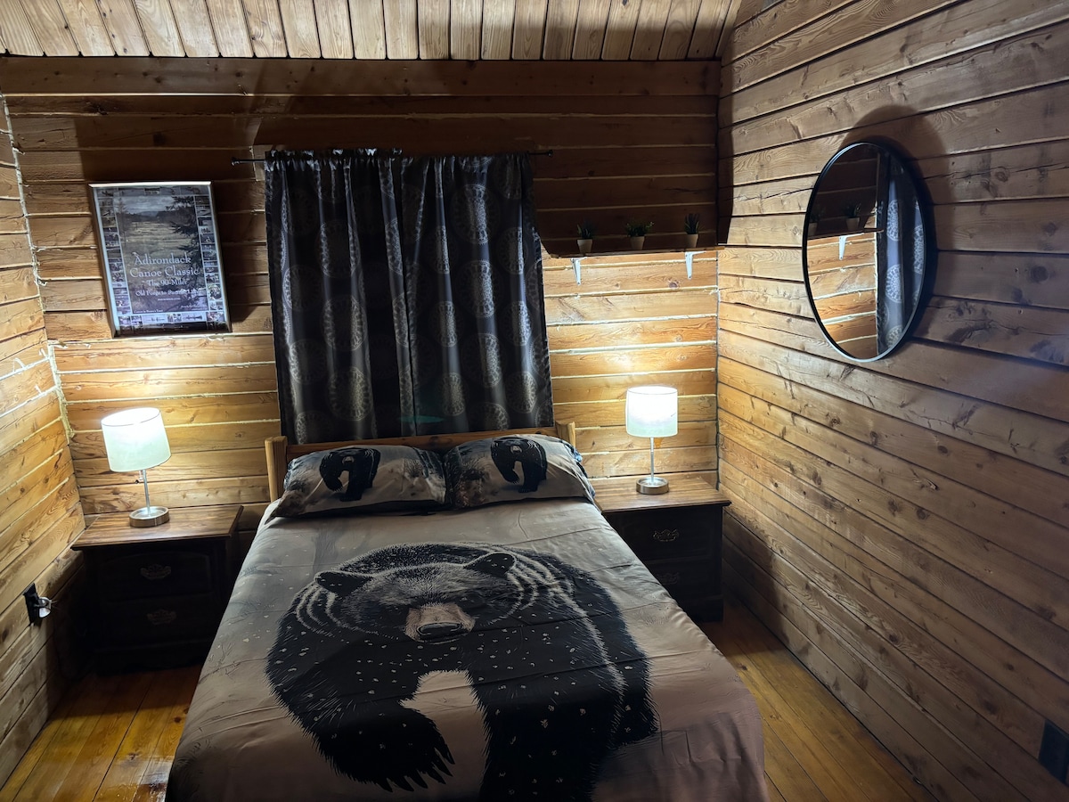 A cozy bedroom features wood-paneled walls and a spacious bed adorned with a bear-themed blanket. Two bedside tables with lamps provide ambient lighting. A large round mirror is mounted on the wall, and a framed print hangs beside a window, partially covered by a dark curtain.