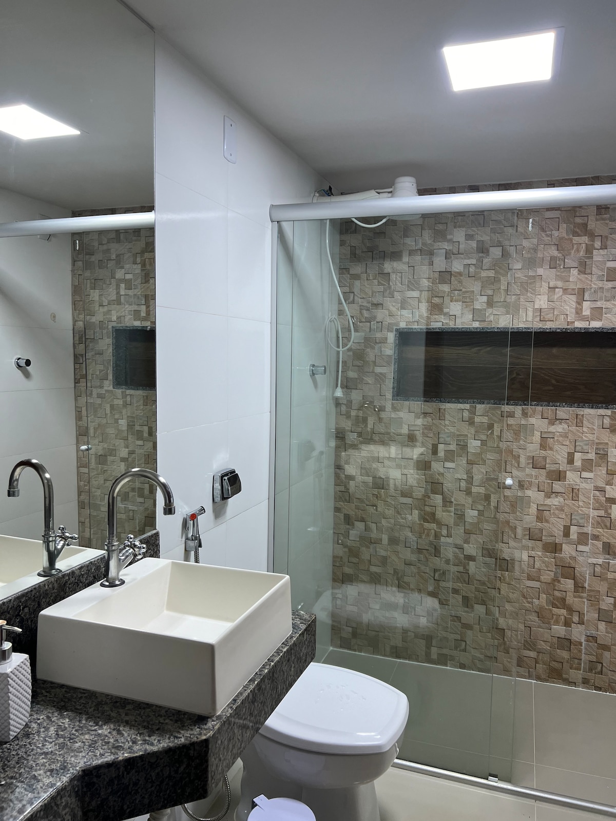 A modern bathroom features a walk-in shower with glass panels, complemented by light tile work on the walls. A stylish basin is positioned on a dark granite counter, with contemporary faucets and a large mirror reflecting the clean design.