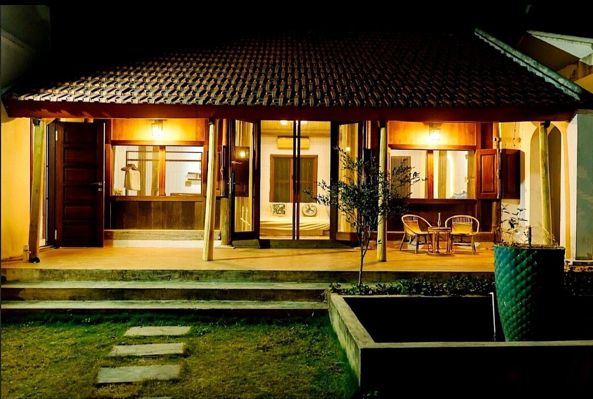 A charming exterior view of the retreat showcases a warm, inviting entrance with a tiled roof. Large glass doors are framed by wooden accents, revealing glimpses of the interior. Soft lighting highlights the outdoor seating area, and a well-maintained garden complements the serene atmosphere.