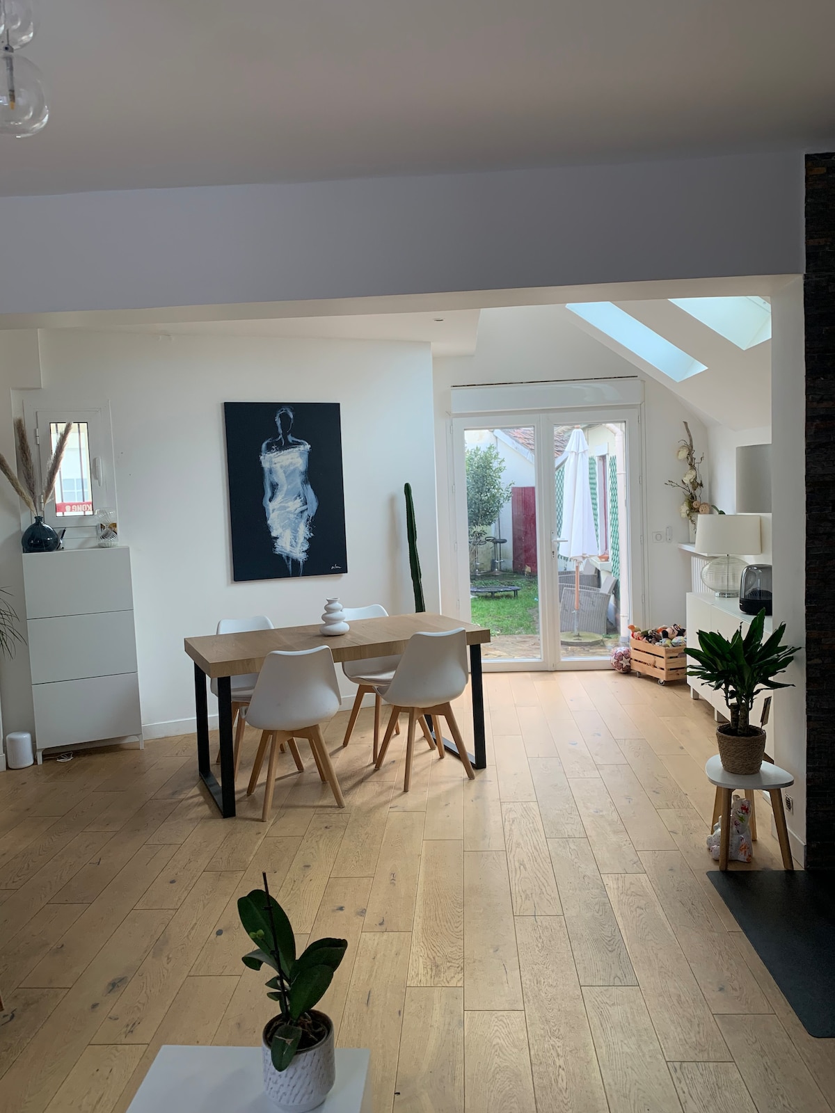 A dining area is showcased with a wooden table and four white chairs. A piece of artwork is hung on the wall, while a window and glass doors provide access to a garden space. Green plants add a natural touch to the interior.