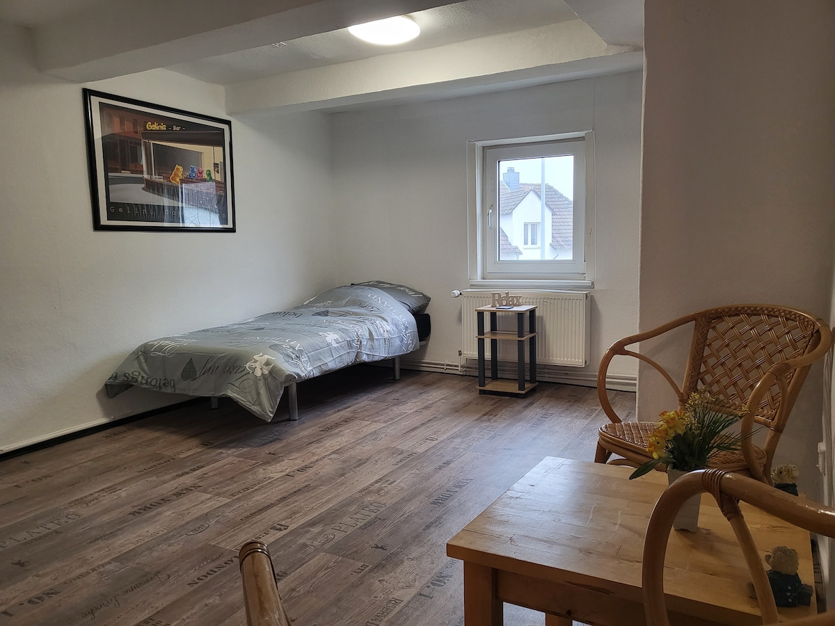 A simple room features a single bed covered with a patterned blanket. A window lets in natural light, and a small wooden table with a chair is positioned nearby. A framed artwork adorns one wall, adding a personal touch to the space.
