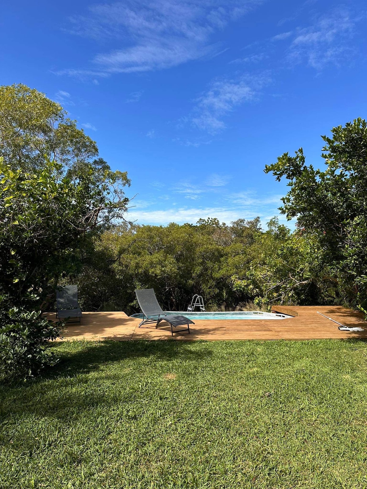 A swimming pool is situated on a wooden deck, surrounded by lush greenery. Sun loungers are positioned nearby, offering a place to relax. The clear blue sky overhead complements the serene outdoor setting.