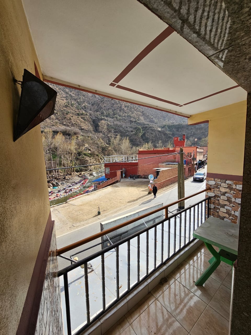 A balcony view is presented, showcasing a mountainous landscape. The scene includes a nearby street, vehicles, and a simple green table. Natural light illuminates the area, enhancing visibility of the surrounding greenery and structures.