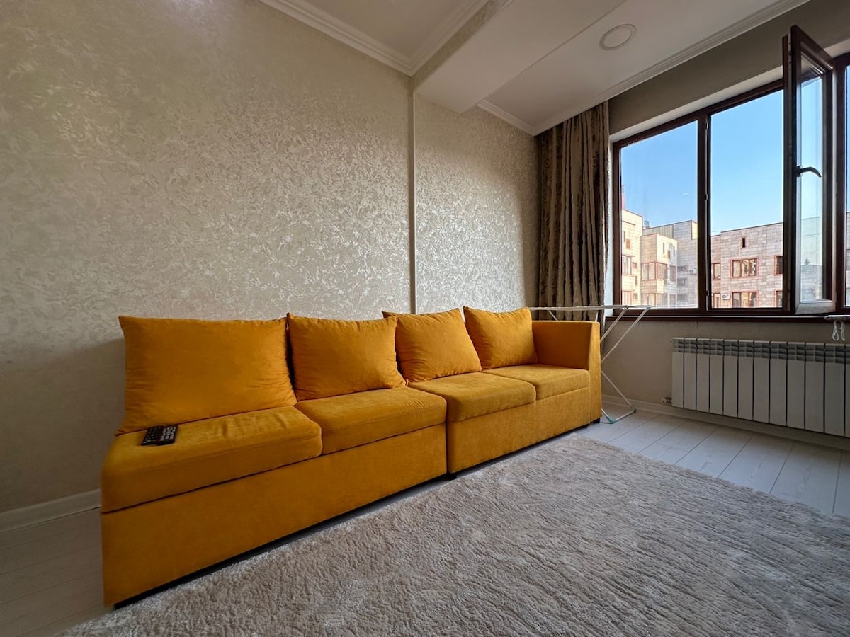 A spacious yellow sofa is positioned against a textured wall, providing ample seating. Large windows adorn one side, allowing natural light to fill the room. A soft area rug lies beneath, adding warmth and comfort to the space.