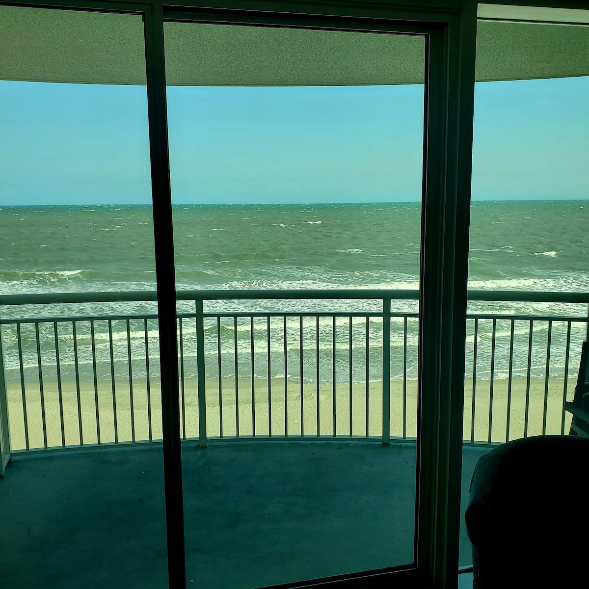 A view of the ocean is captured through expansive glass doors, revealing gentle waves lapping against the sandy beach. The balcony features a white railing, framing the tranquil coastal scene with clear skies above.