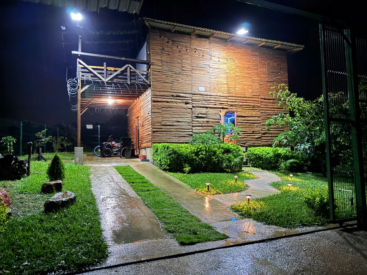 The exterior of a two-story wooden house is viewed at night, with soft lighting illuminating the pathways and garden. Rain can be seen falling gently, while the house features a spacious porch and large windows. Lush greenery lines the walkway, enhancing the natural environment.