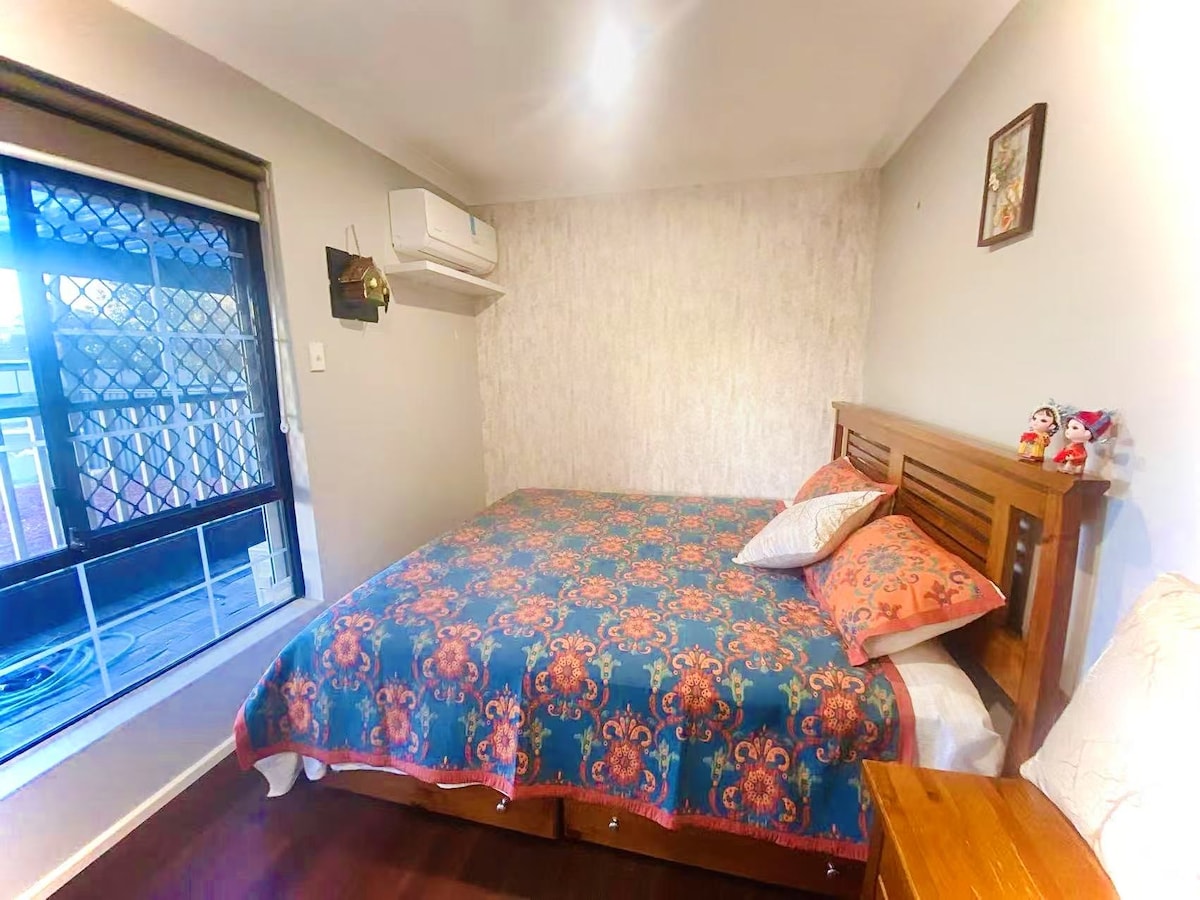 A comfortable bedroom features a spacious bed adorned with a colorful patterned blanket. Decorative pillows are arranged on the bed. A window with a mesh screen allows natural light, while a wall-mounted air conditioning unit is present, contributing to a pleasant indoor environment.