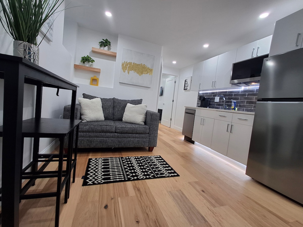 A modern living space is highlighted by a cozy gray sofa adorned with two white cushions. A black and white patterned rug sits beneath a small black side table. The kitchen is equipped with stainless steel appliances and features light cabinetry against a dark tiled backsplash.