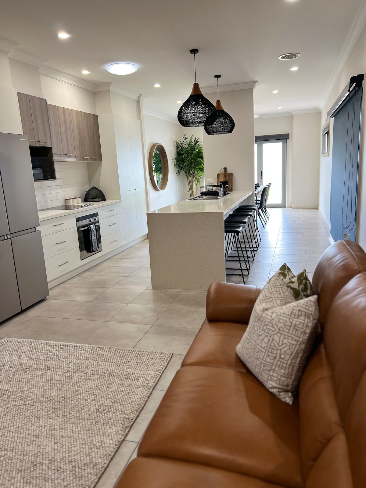 The kitchen area features modern appliances and a spacious island with seating. Stylish pendant lights hang above, and a circular mirror reflects the light. A comfortable sofa with a decorative pillow is situated nearby, with a textured rug underfoot and a potted plant adding a touch of greenery.