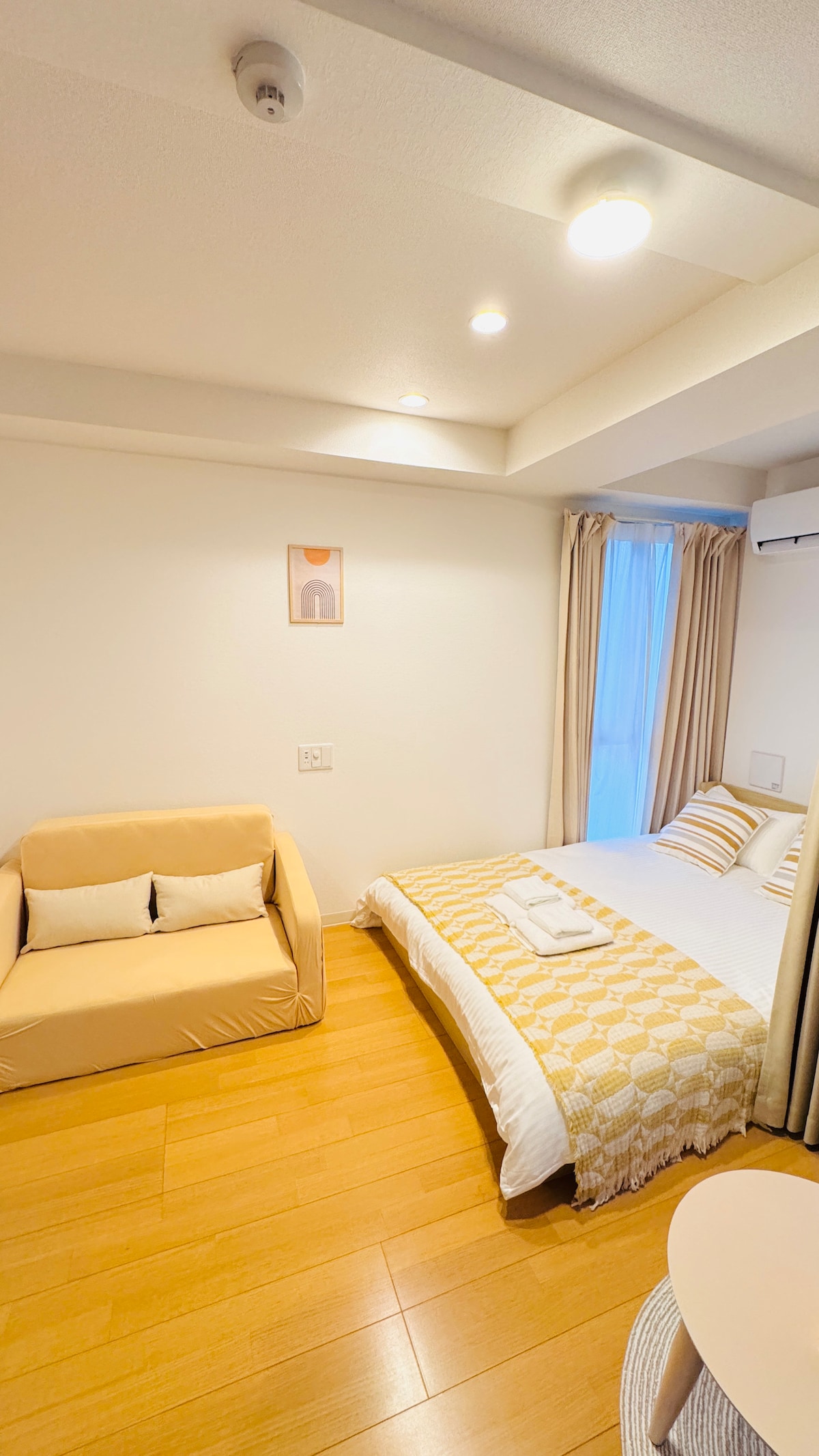 A cozy room features a spacious bed dressed in soft linens, complemented by a light blanket. A small sofa is placed nearby, creating a relaxing seating area. Natural light filters through the partially draped window, illuminating the warm wooden floor.
