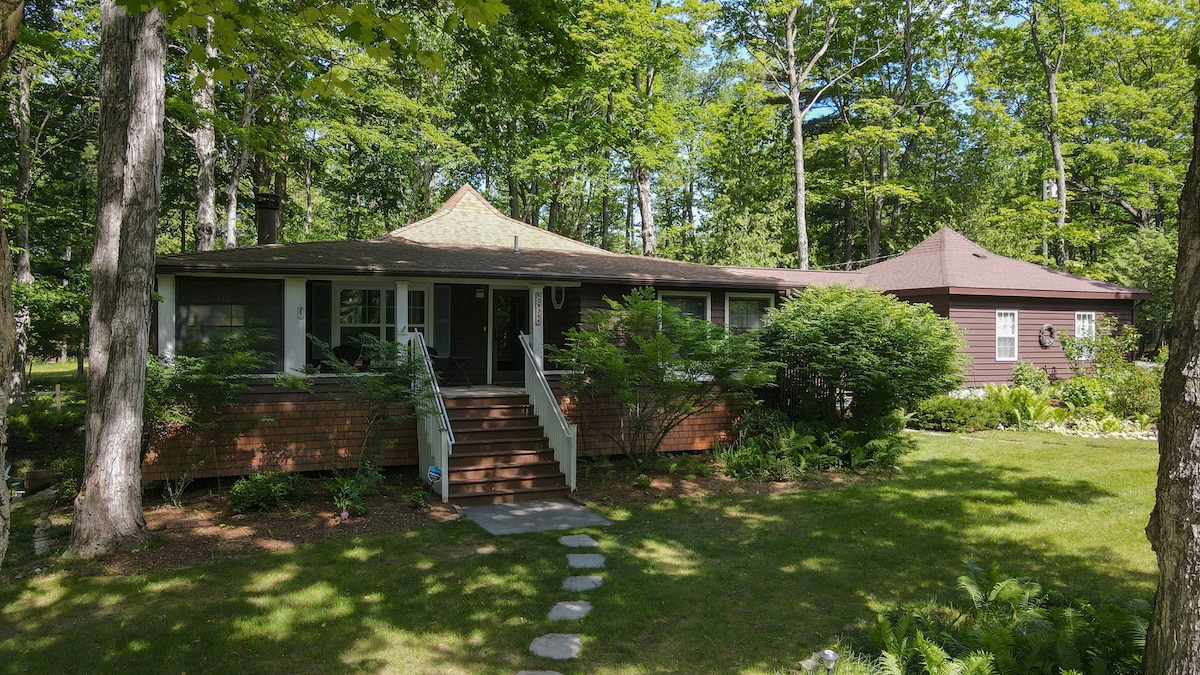 A cabin surrounded by lush greenery features a welcoming entrance with wooden steps leading to a spacious porch. Large windows enhance the view of the landscaped yard, which includes a path of stepping stones. The structure blends harmoniously with the natural environment.