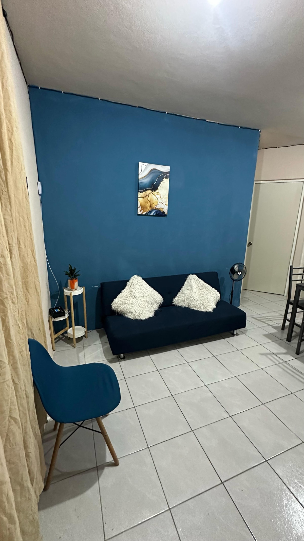 A compact living space features a dark blue accent wall, complemented by a comfortable sofa with decorative pillows. A small round table with a plant stands nearby. Light-colored curtains frame the window, while a fan is positioned on the floor across from the seating area.