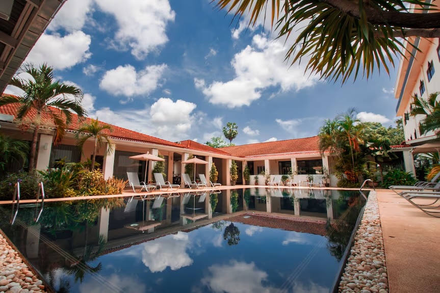 A serene swimming pool is surrounded by well-maintained gardens and palm trees. Lounge chairs are positioned nearby for relaxation. The pool's surface reflects the blue sky dotted with white clouds, enhancing the tranquil setting of the hotel.