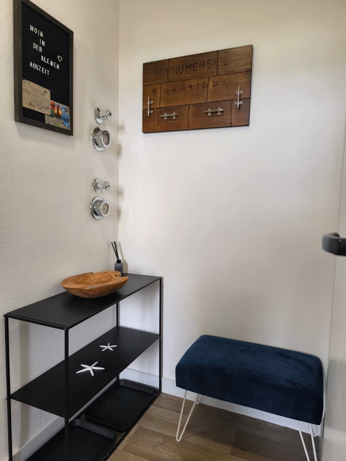 An inviting entryway showcases a black shelving unit displaying decorative elements. A wooden bowl and a decorative sign titled 'Dornumersiel' add charm. Light-reflecting mirrors are mounted on the wall, and a blue velvet stool provides a comfortable seating option.