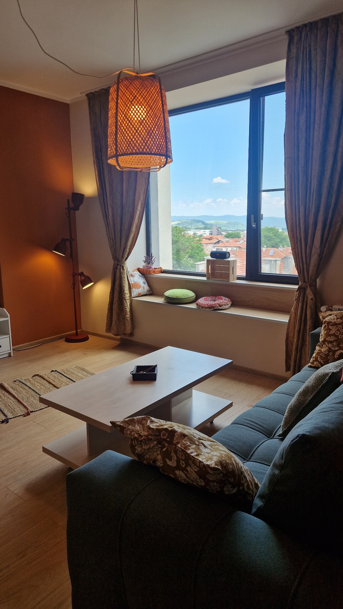 A cozy living area features a comfortable dark green sofa adorned with decorative cushions. A light wooden coffee table is positioned in front, with a lamp illuminating the space. A large window frames a scenic view, allowing natural light to brighten the room.