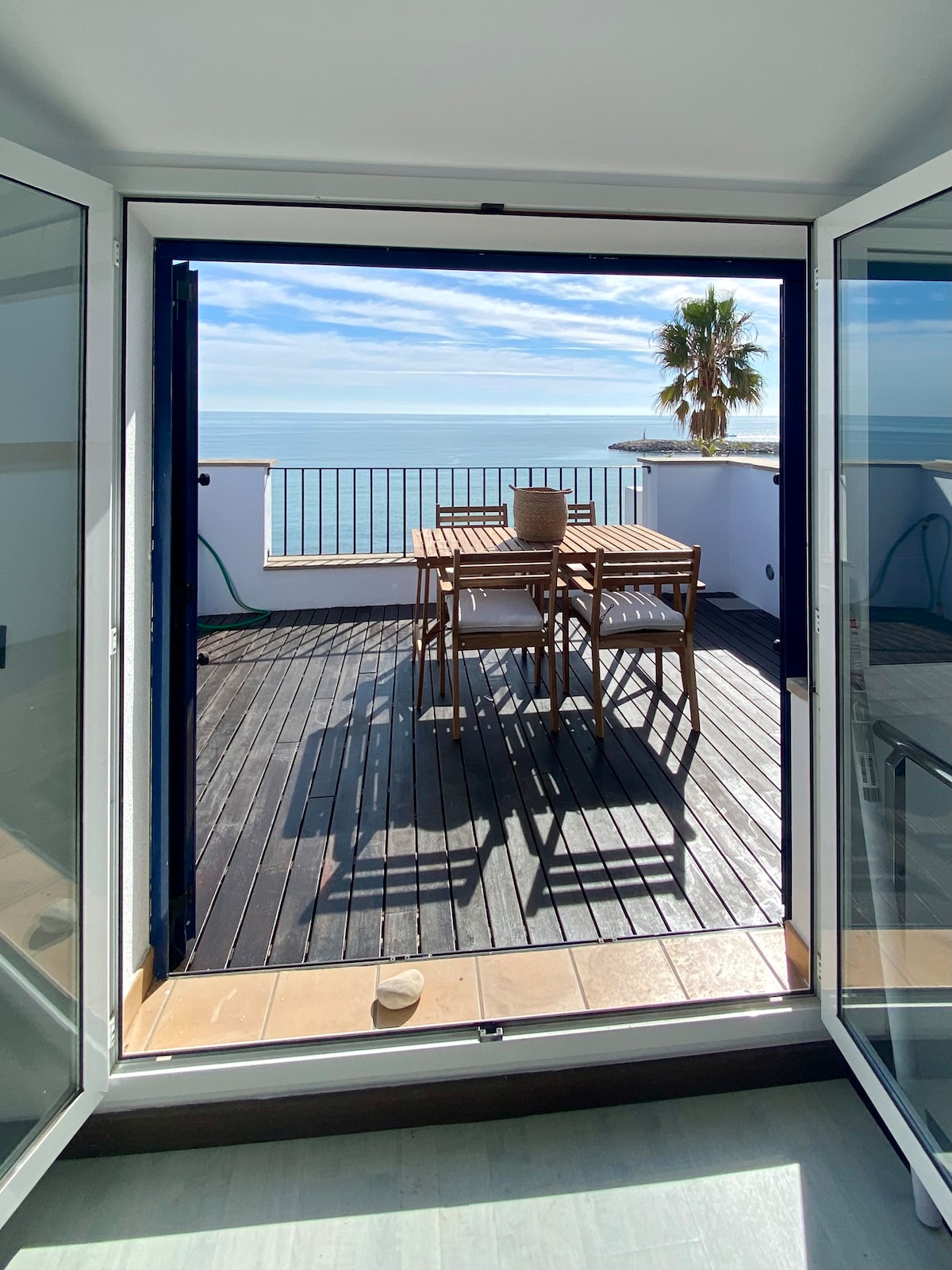 A spacious terrace opens up from a set of glass doors, featuring a wooden dining table accompanied by four chairs. The serene sea can be seen in the distance, with sunlight reflecting off the water, creating a calm atmosphere.
