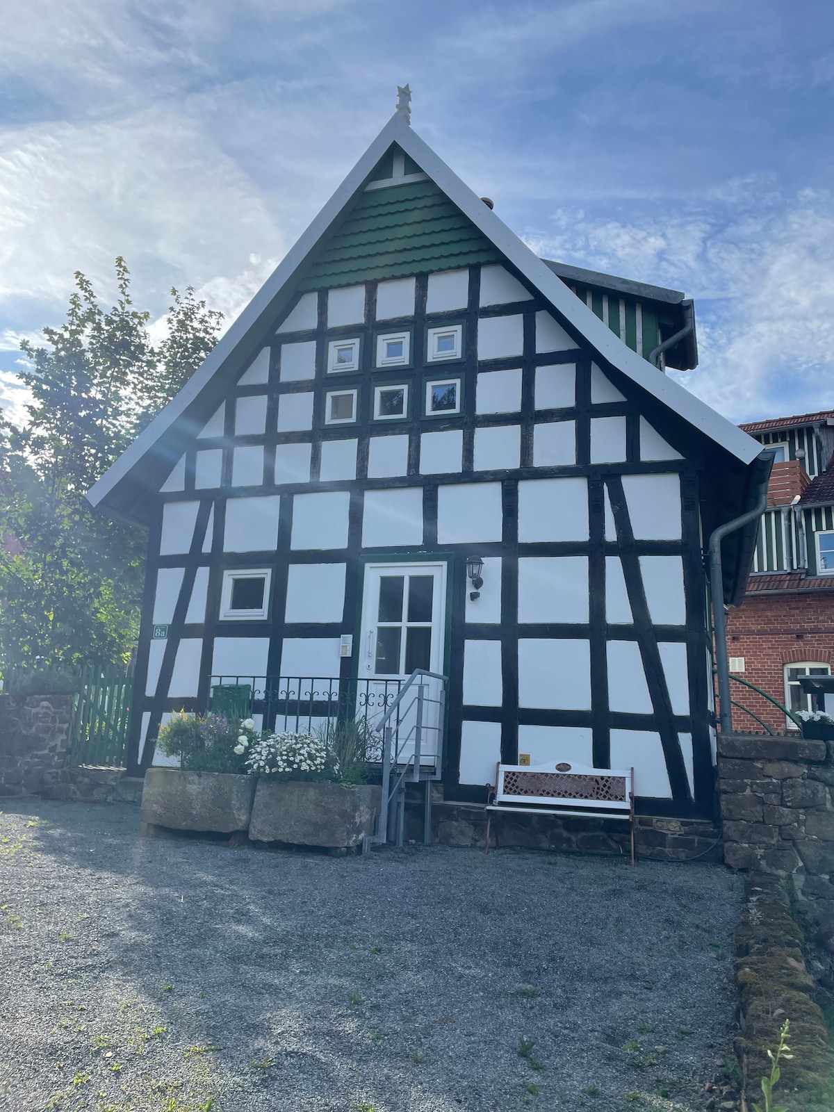 The exterior of a charming half-timbered house showcases a mix of white and dark wood elements. A gravel pathway leads to the entrance, where stairs are visible. Surrounding greenery adds to the tranquil setting, enhancing the inviting appearance of this unique structure.
