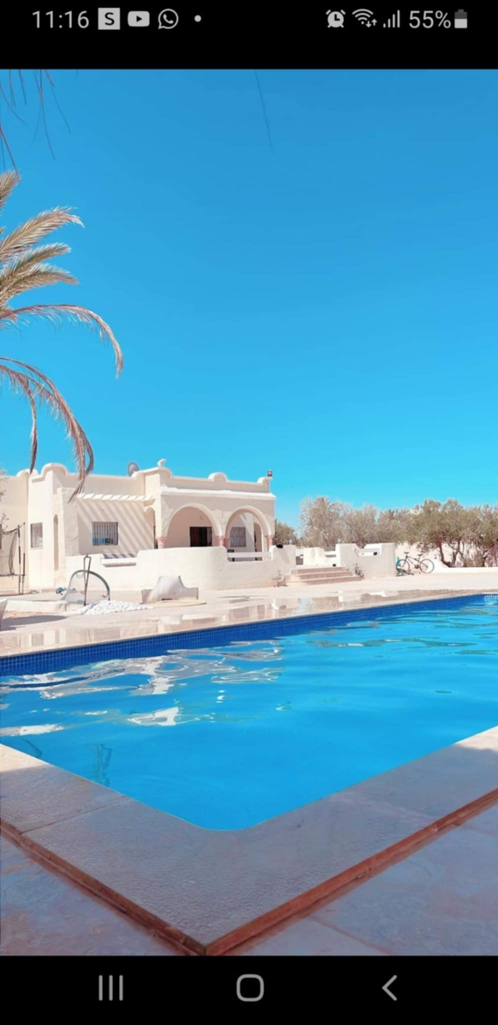 A spacious outdoor area features a clear blue swimming pool with steps leading down into the water. The villa, with its light-colored stucco exterior, is set against a bright, clear sky. Palm trees provide greenery around the swimming area.