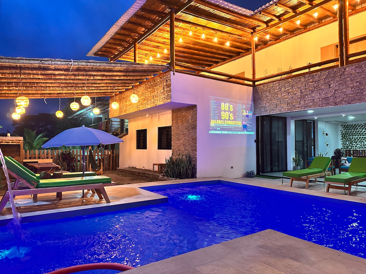 A vibrant outdoor space features a large swimming pool surrounded by lounge chairs. Soft lighting creates a relaxing atmosphere. A patio area is designed with a seating arrangement, and a parasol offers shade. The two-story structure showcases glass doors, enhancing the inviting setting.
