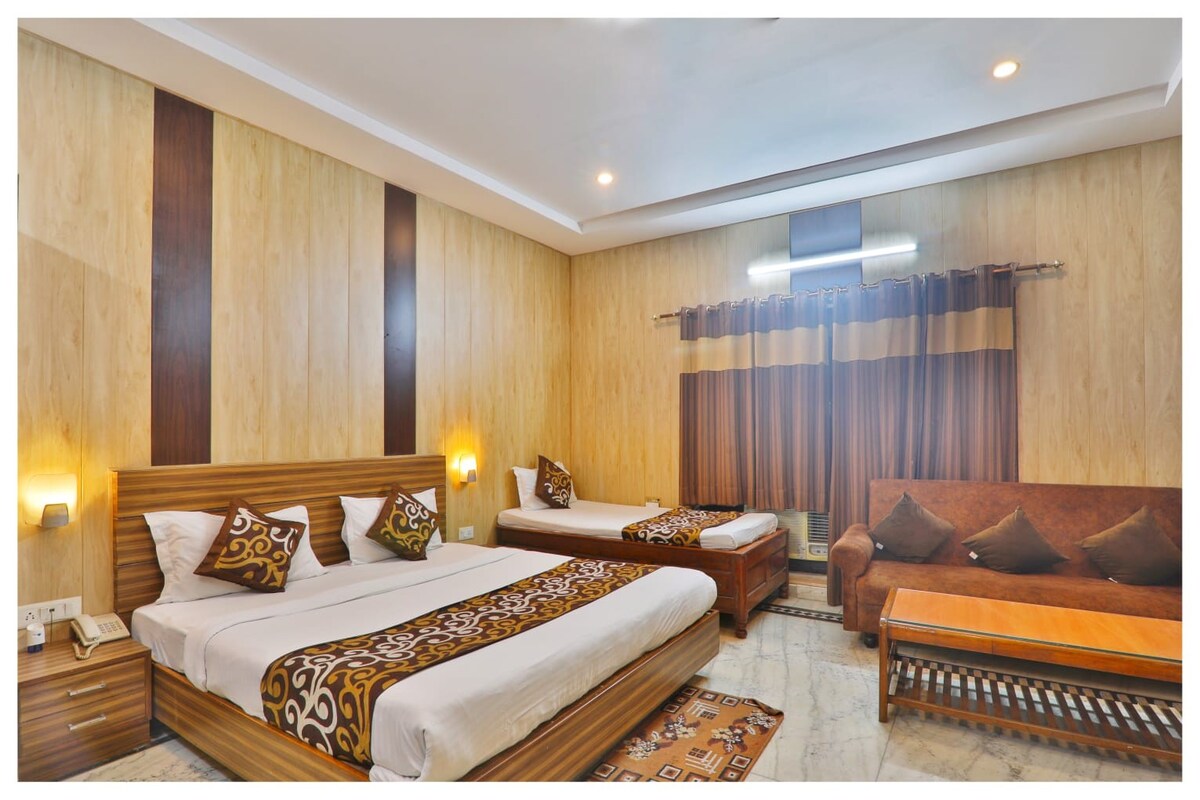 A spacious guest room features two beds with decorative brown and gold fabric accents. A comfortable sofa is positioned near the wall, accompanied by a wooden coffee table. Natural light filters through large curtains, illuminating the warm wood paneling throughout the space.
