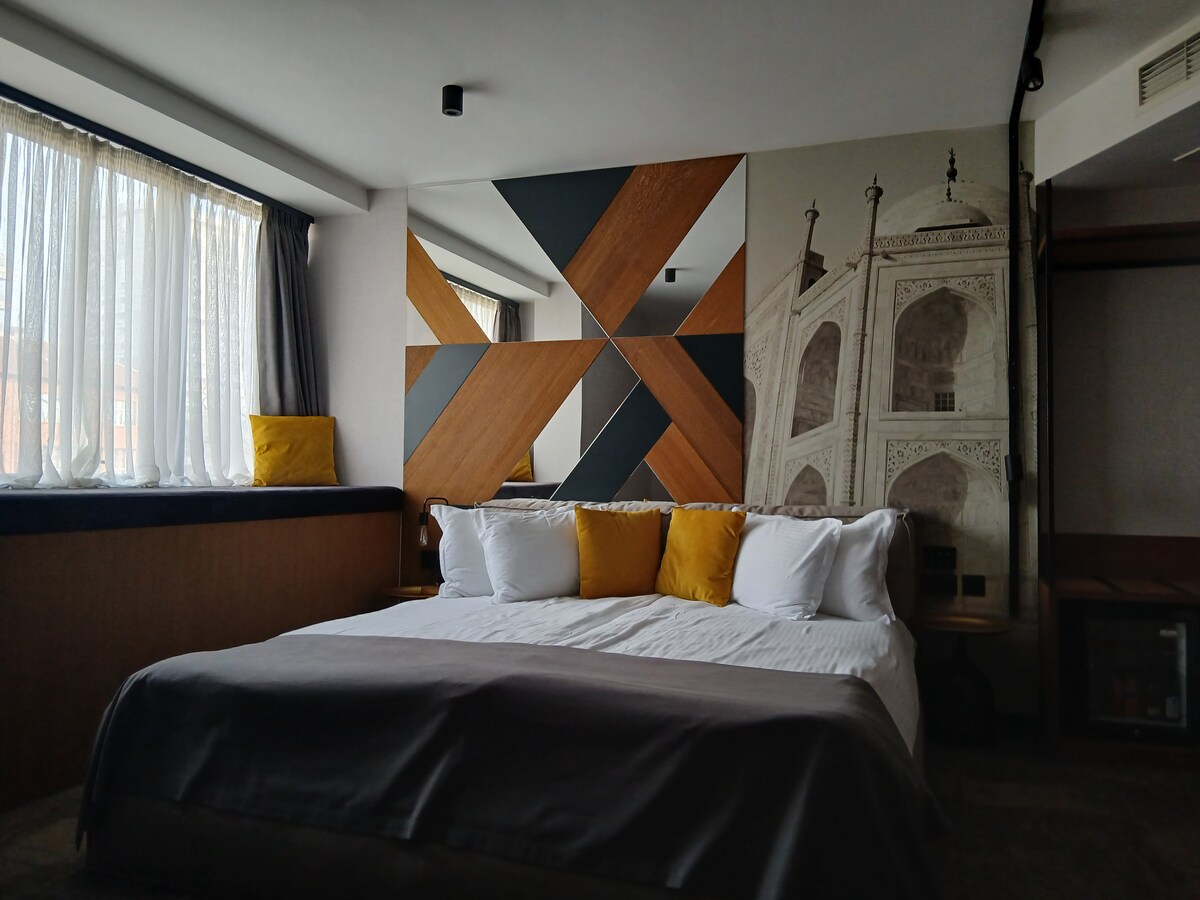 A spacious bedroom is highlighted by a large bed adorned with soft white linens and cozy gray blankets. Geometric wood accents and a mural depicting an architectural feature complement the minimalist decor. Natural light filters through sheer curtains, adding warmth to the inviting atmosphere.