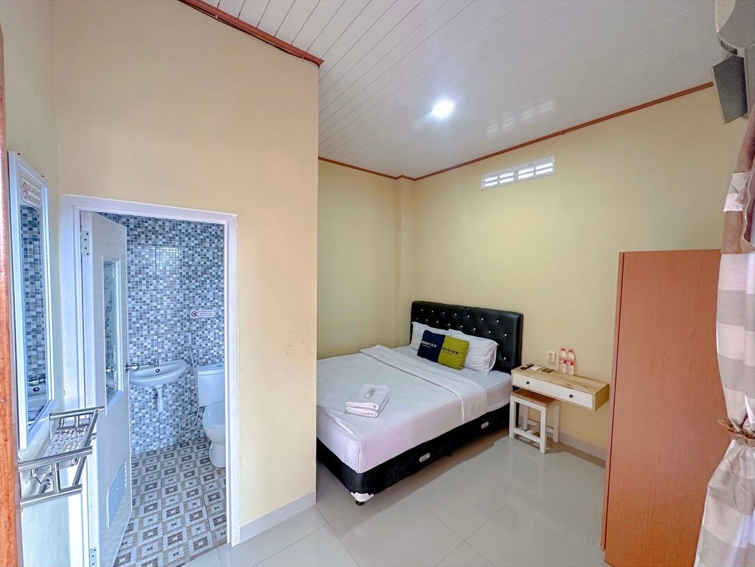 A spacious room features a large bed with a padded black headboard, complemented by a side table. A bathroom with modern tiles is accessed through a door, while large windows allow natural light to brighten the softly colored walls.