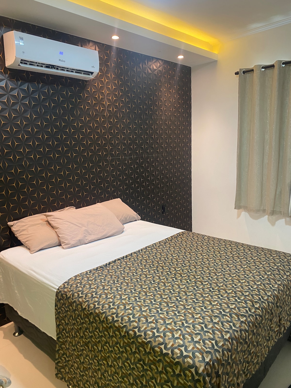 A cozy sleeping area features a spacious bed covered with a patterned blanket. A dark geometric accent wall adds visual interest, while warm lighting is provided by recessed fixtures above. The air conditioning unit is mounted on the wall, and neutral curtains frame the window.
