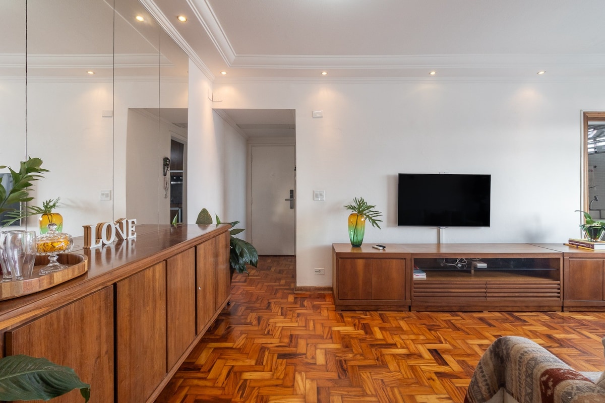 The living area features wooden flooring and a stylish console table adorned with decorative items. A flat-screen TV is mounted on the wall, beside a large window that lets in natural light. Indoor plants add a touch of greenery to the space.