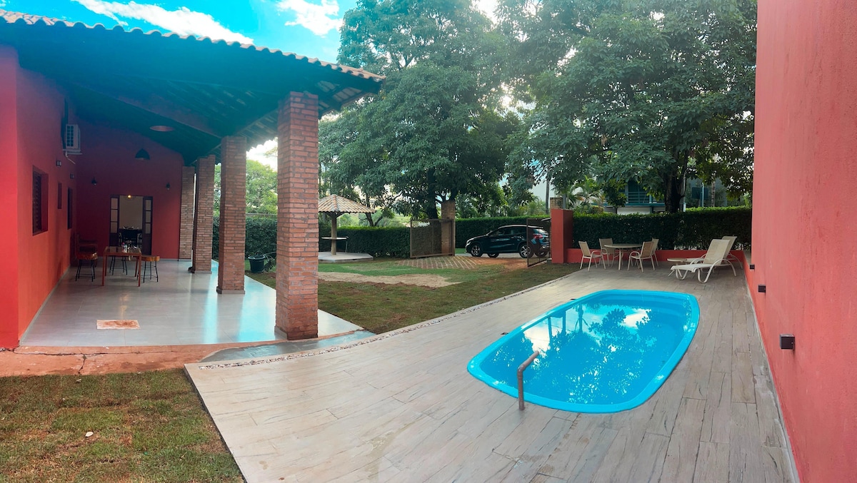 An outdoor area is shown featuring an inviting pool surrounded by a tiled deck. Nearby, lounge chairs offer additional seating options. Lush greenery and trees create a serene atmosphere, with a covered patio visible in the background, providing shade and space for gatherings.