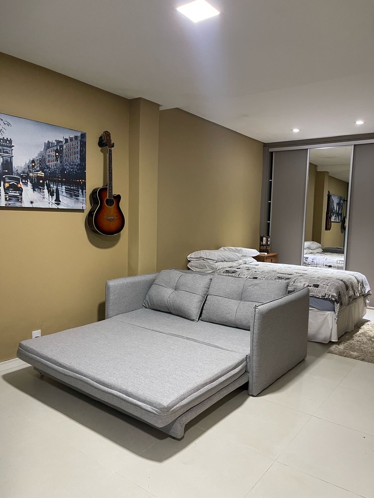 A spacious area features a gray sofa bed, positioned near a wall adorned with a framed artwork depicting a city scene. An acoustic guitar hangs on the wall, while a mirrored wardrobe reflects the cozy sleeping space, which includes a neatly made bed with linens.