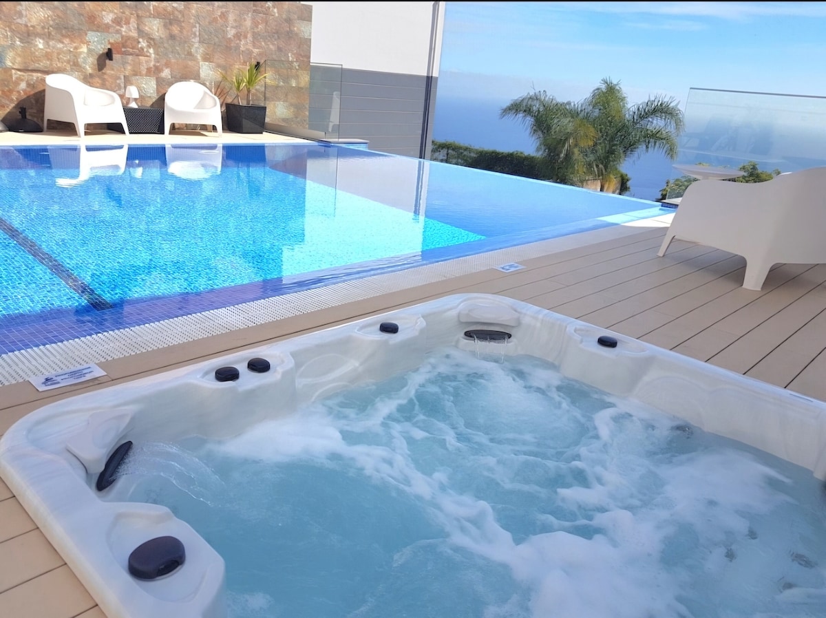 A shared area features a white jacuzzi with bubbling water and adjacent shimmering pool. The surrounding deck is lined with modern lounge chairs, and a panoramic view of the sky and distant hills is visible in the background.