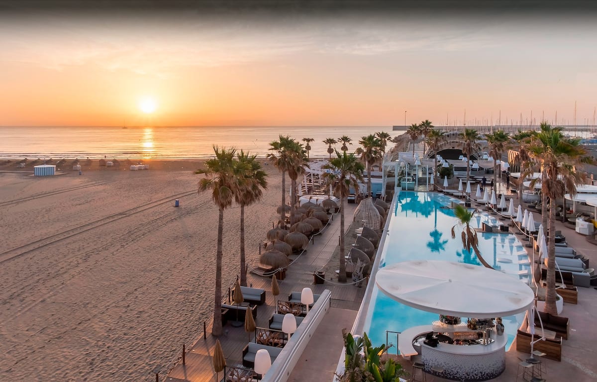 A serene beachfront scene captures the sun rising over the ocean, with gentle waves lapping at the shore. A stylish pool is seen alongside the sandy beach, bordered by palm trees and lounge chairs. Umbrellas and beachside facilities are also visible, creating a relaxed atmosphere.