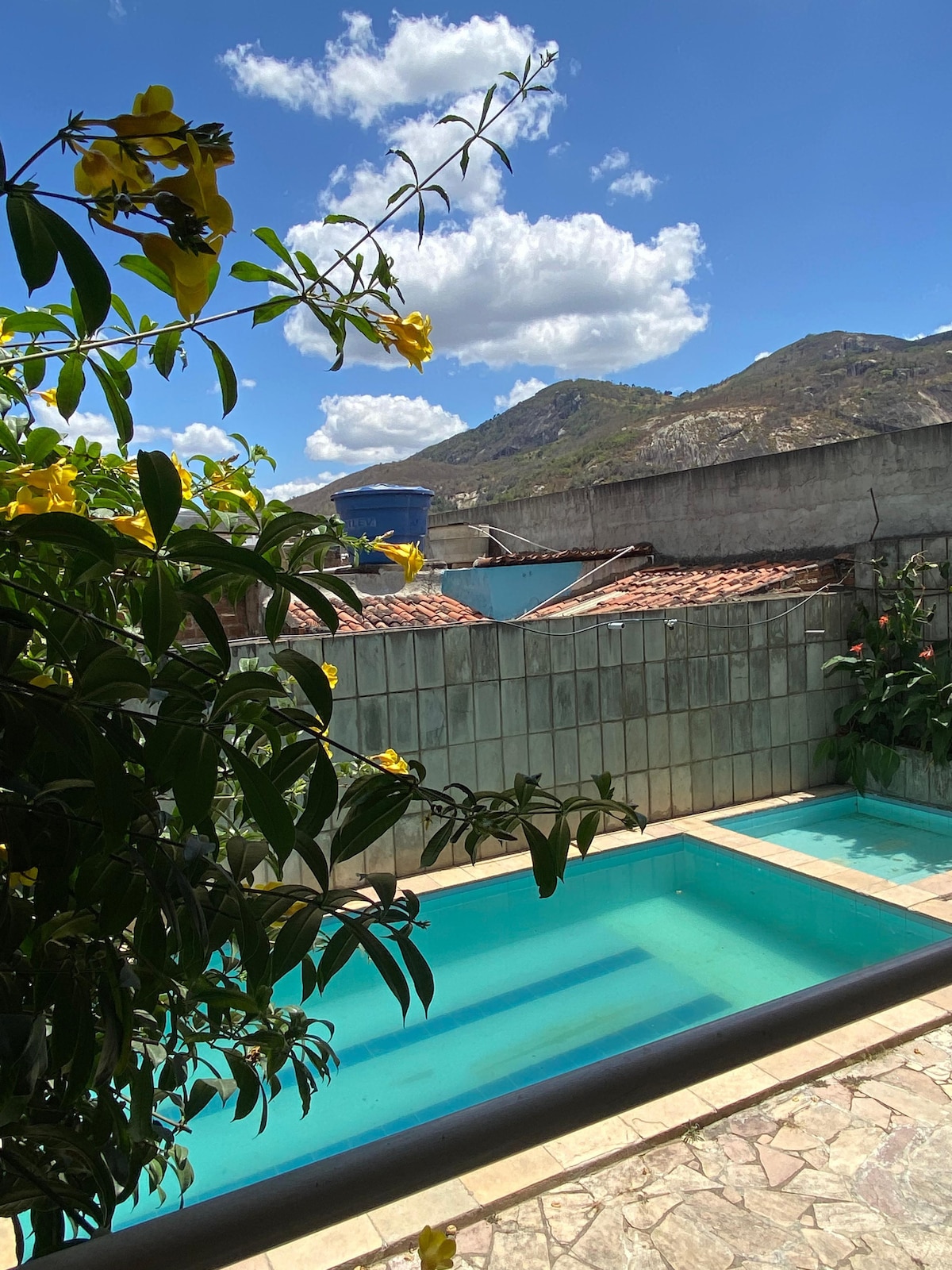 A view of two serene swimming pools is framed by yellow flowers and greenery. Behind, a backdrop of mountains is visible against a clear blue sky with scattered clouds. The area features a combination of textured stone and tiled flooring.