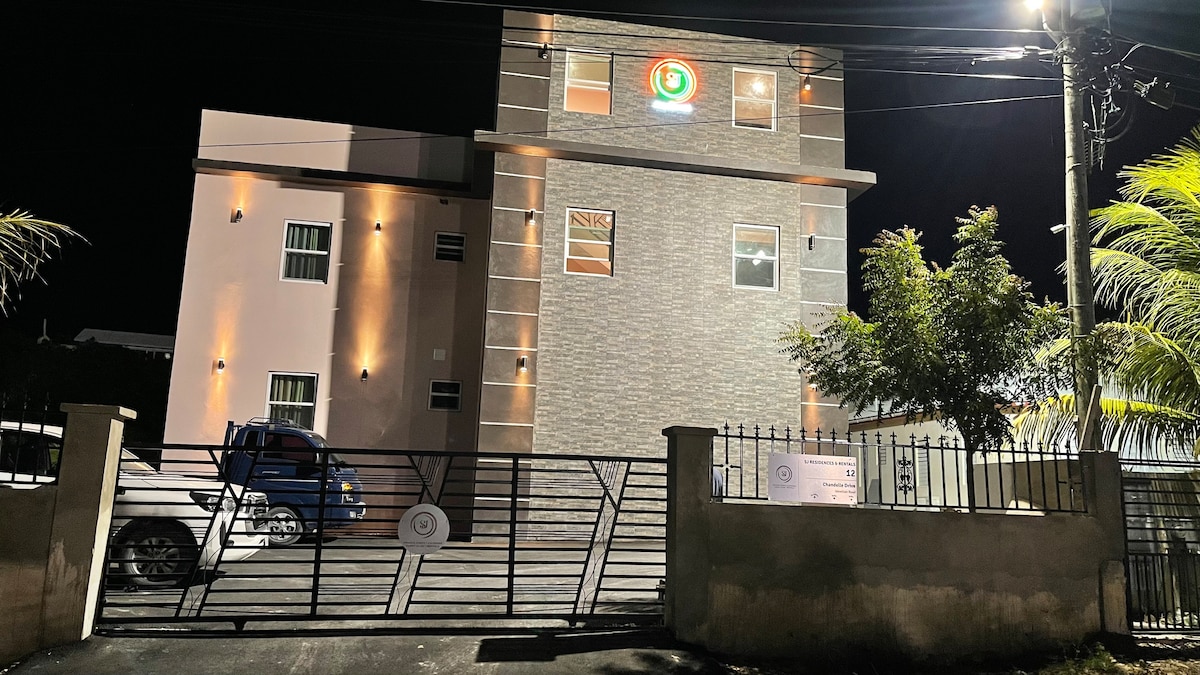 The exterior of the SJ Residences is captured at night, showcasing a modern three-story building with stone accents and illuminated signage. A gated entrance provides access to a driveway, where parked vehicles are visible under well-lit surroundings.