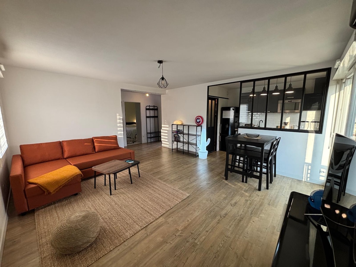 The spacious living area is furnished with an orange sofa, coffee table, and a woven rug. A dining set with black wooden chairs is positioned near a window. The kitchen is visible in the background, featuring sleek cabinetry and modern appliances.