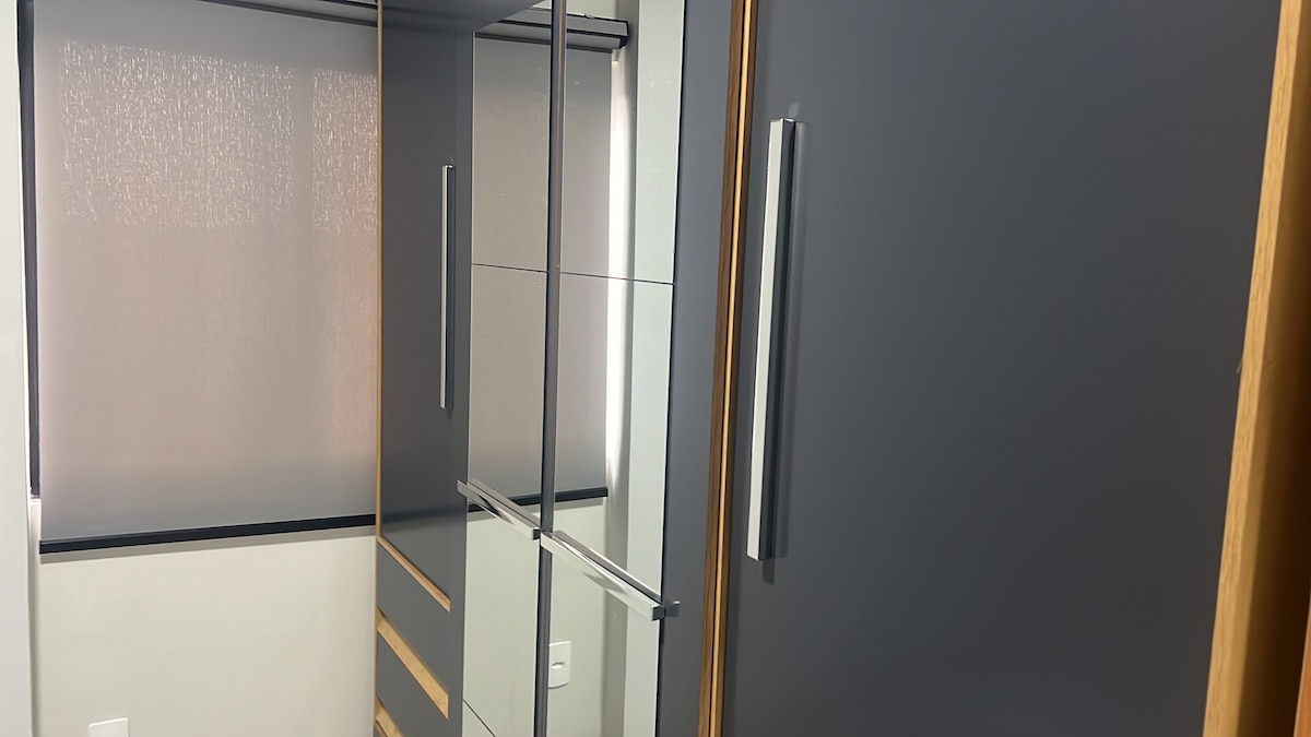 A modern wardrobe with mirrored doors reflects the interior space. The cabinetry features a sleek design with wooden accents and a smooth finish. Natural light filters gently through a light-colored window covering, enhancing the contemporary feel of the room.