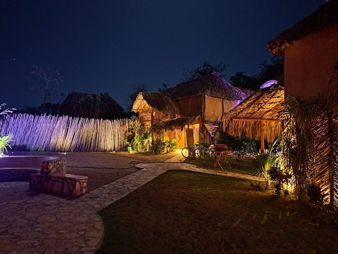 Traditional thatched-roof cabins are illuminated under the night sky, surrounded by soft landscaping and a stone pathway. The area is accented with warm lighting, enhancing the natural textures of the structures and the greenery nearby.