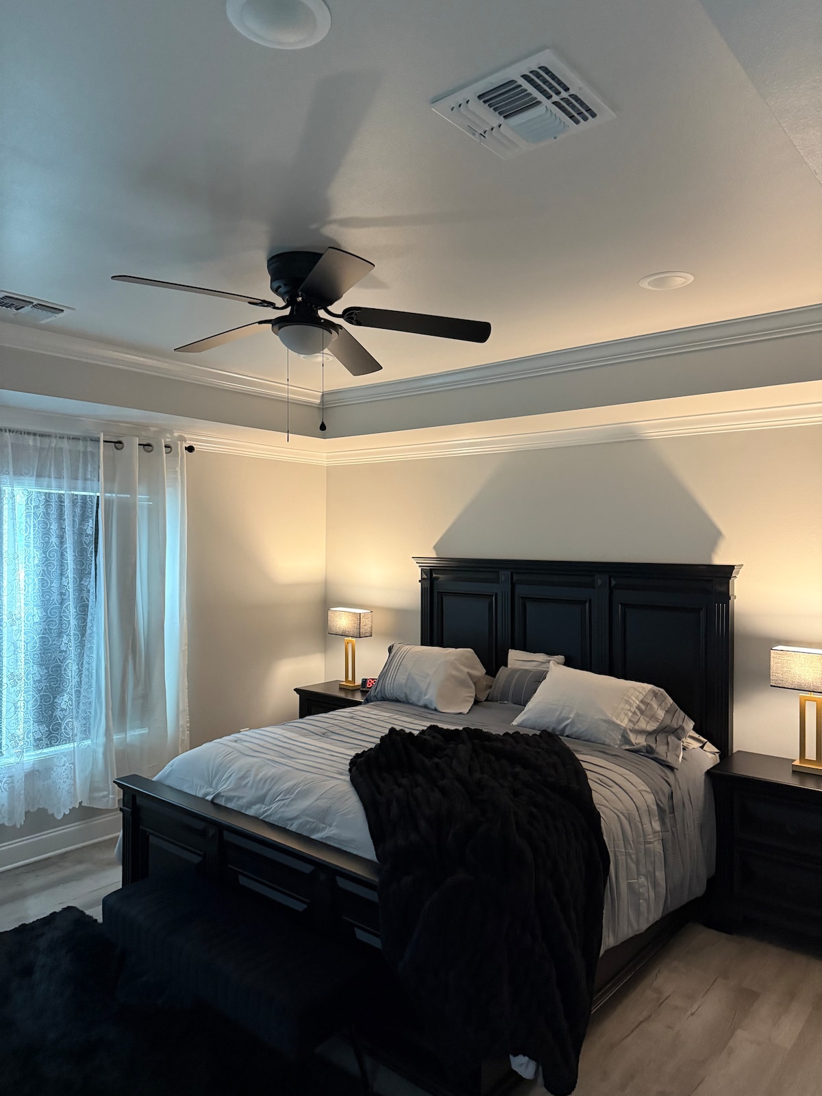 A serene bedroom showcases a dark wood headboard and matching nightstands. The spacious bed is neatly dressed with white linens and flanked by two lamps. A soft, textured blanket is draped across the foot of the bed. Natural light filters through a sheer-curtained window, emphasizing the room's calm ambiance.