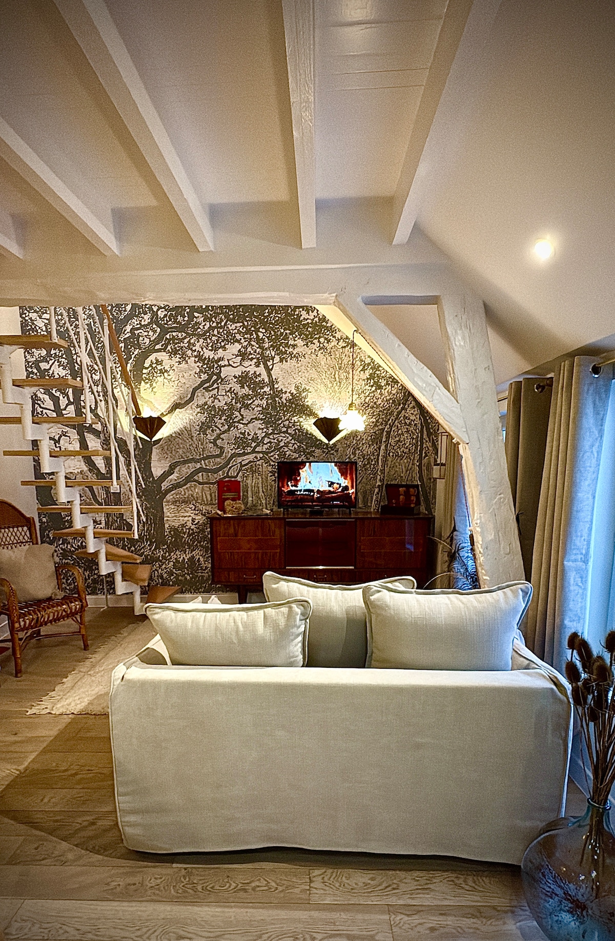 A cozy living area features a light-colored sofa centered around a wooden entertainment unit. A mural depicting a serene landscape adorns the wall, while elegant pendant lights provide soft illumination. A staircase with wooden railings leads to the upper level, enhancing the space's charm.