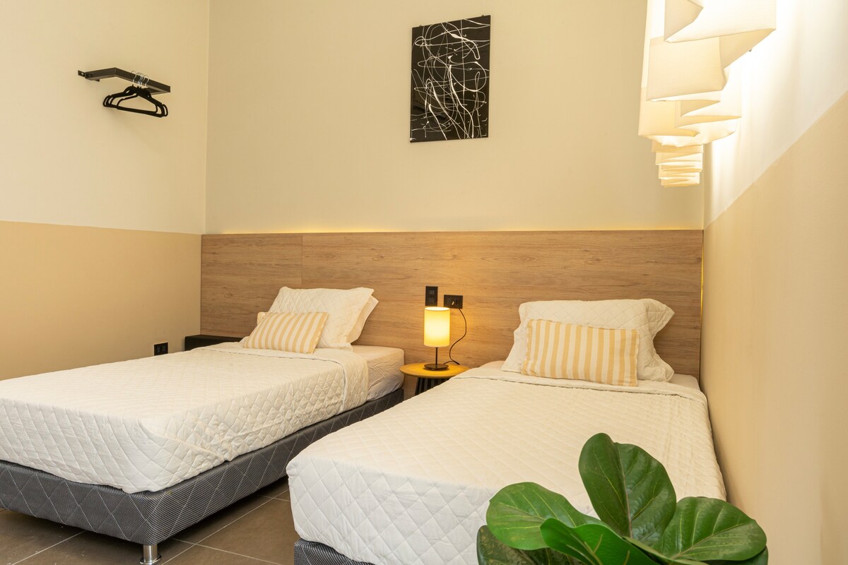 The room features two individual beds with quality bedding and striped pillows. A small nightstand with a lamp is positioned between the beds. A minimalist wall art piece is displayed above, and a hanger is mounted on the side wall. Natural light enters through an unobstructed window.