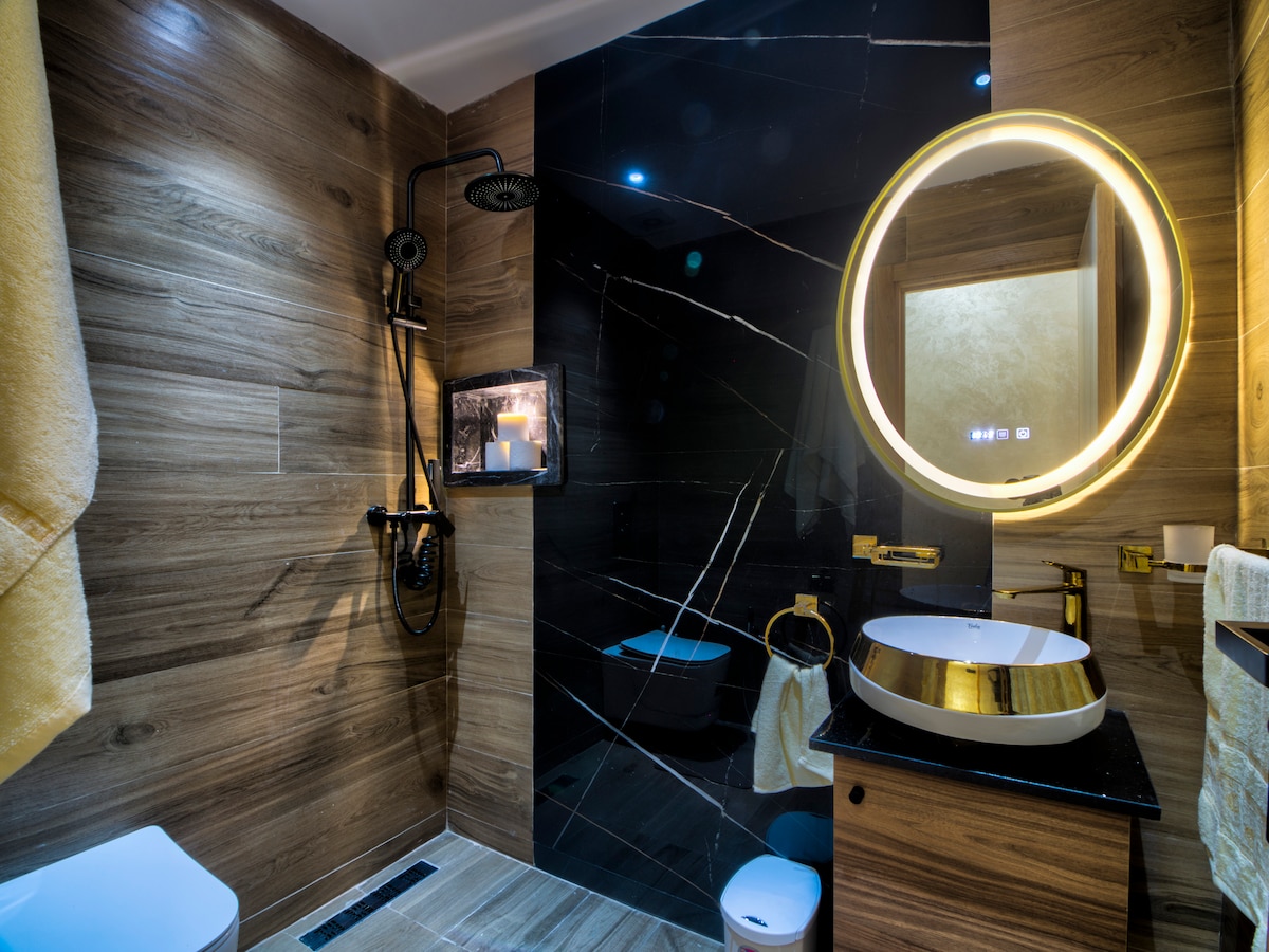 The modern bathroom features a sleek design with dark marble accents and wooden elements. A round backlit mirror is mounted above a stylish sink, while a spacious shower area is equipped with a rain showerhead. Neutral-colored towels are neatly arranged nearby.