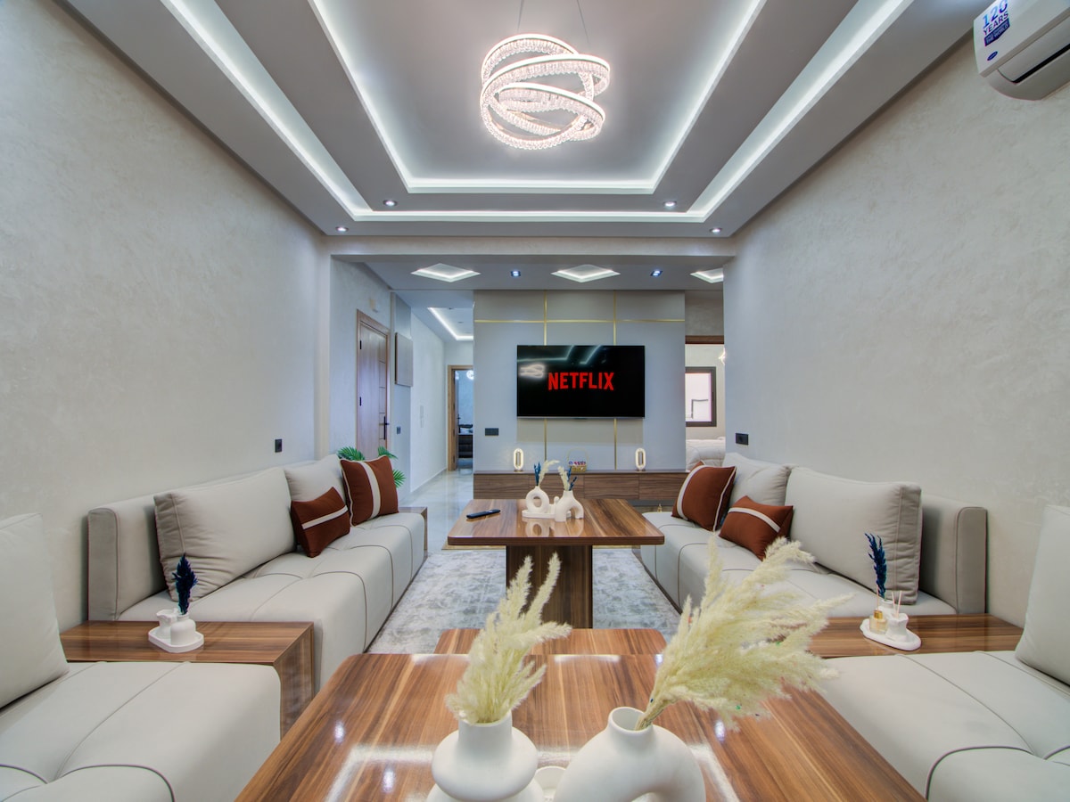 A modern and spacious living area is highlighted by a sleek ceiling with recessed lighting. Soft, neutral-colored sofas are arranged around a wooden coffee table, complemented by decorative accessories. A large television is mounted on the wall, displaying the Netflix logo, enhancing the entertainment options.
