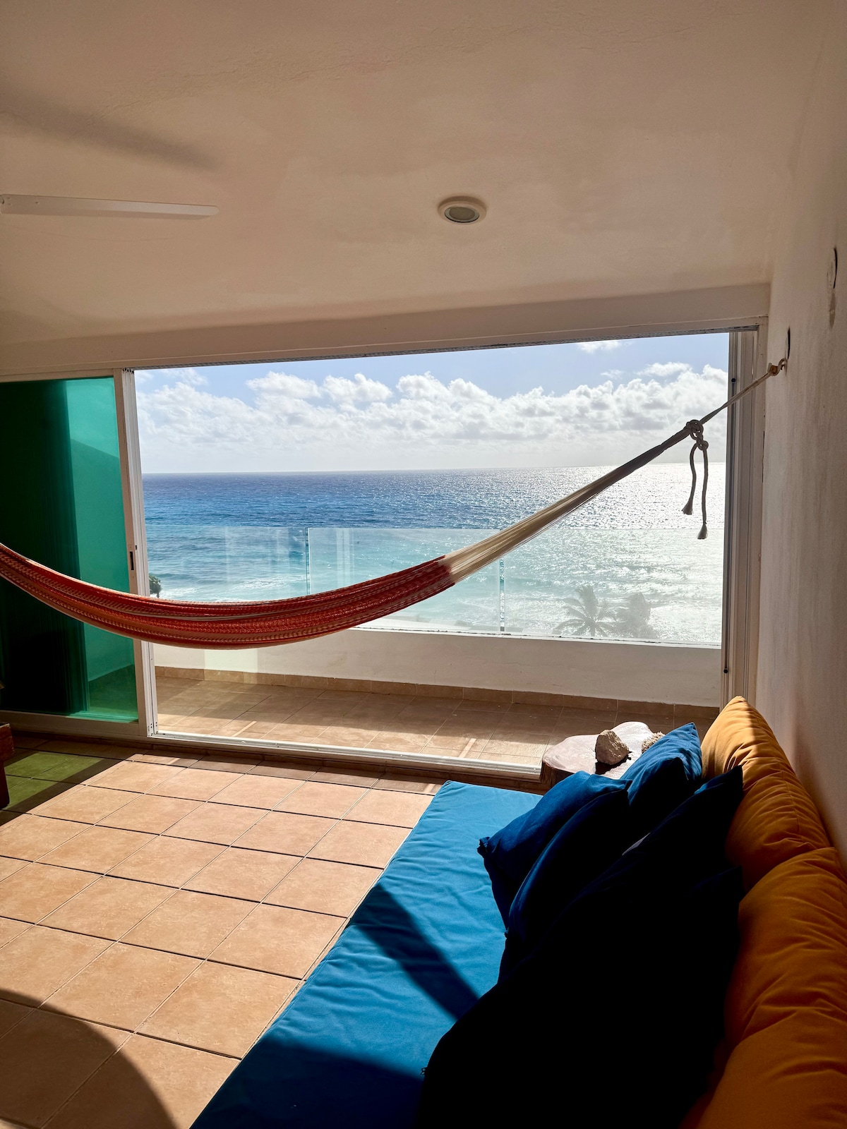 A cozy seating area is highlighted by a colorful couch and a red hammock, positioned near large glass doors. The view showcases the ocean with distant clouds reflecting the sunlight, creating a serene coastal atmosphere.