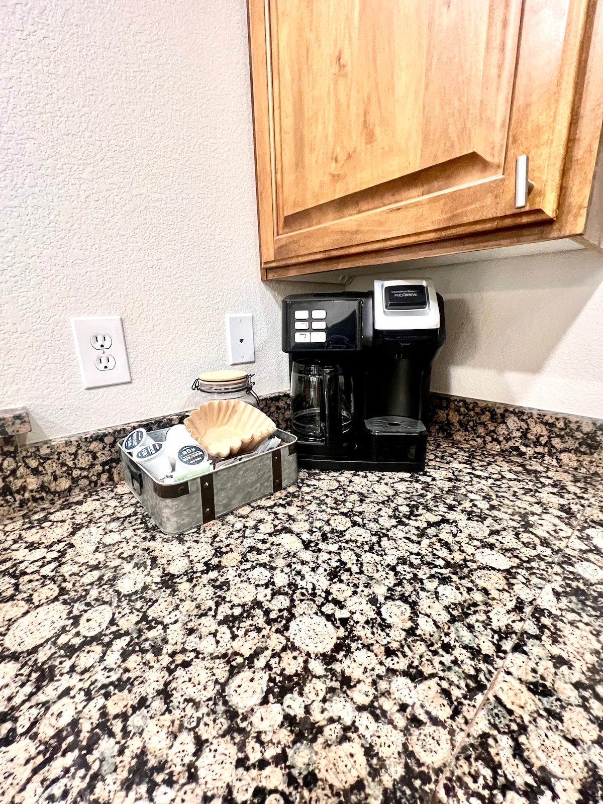 A contemporary coffee maker is positioned on a speckled granite countertop, along with a small metal container holding coffee pods and a decorative shell. Warm wooden cabinets provide contrast above the countertop, enhancing the functional kitchen space.