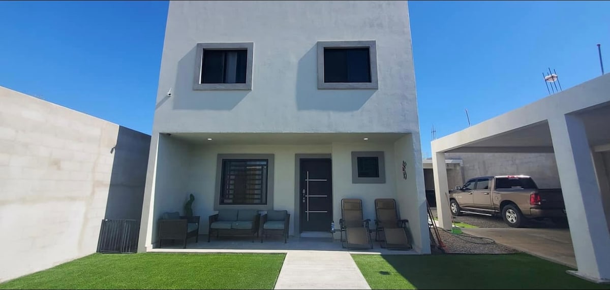 The exterior of a modern two-story house is displayed, featuring a covered porch with comfortable seating arranged on the left. A well-maintained lawn surrounds the entrance, and the bright blue sky is visible above, enhancing the clean lines of the architecture.